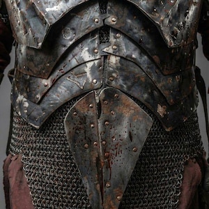 May include: Close-up of a medieval-style chest armor. The armor is dark gray with riveted plates and chainmail. Splatters of red are visible on the metal. The Knights In logo is in the upper left corner.
