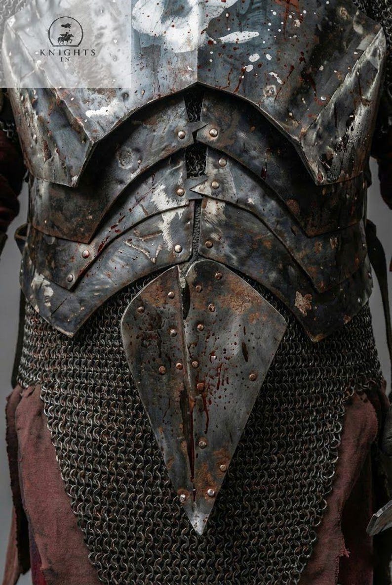 May include: Close-up of a medieval-style chest armor. The armor is dark gray with riveted plates and chainmail. Splatters of red are visible on the metal. The Knights In logo is in the upper left corner.