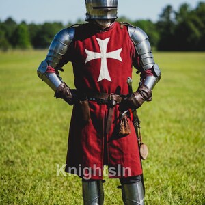 May include: A full suit of medieval armor, including a helmet, chest plate, and leg guards. The red tunic features a white cross. The image includes the text "Knights In". The knight stands in a grassy field.