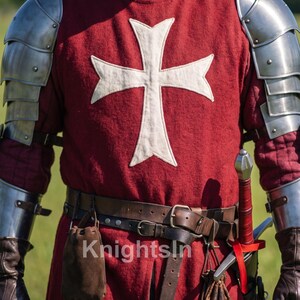 May include: A medieval knight's costume featuring a red tunic with a white cross emblem, metal armor, and a leather belt with pouches. The costume includes a sword and is labeled "Knights In".