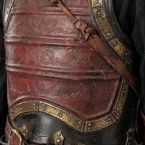 May include: Close-up of a medieval-style leather breastplate with intricate embossed designs. The armor features a deep red central panel, accented with brass-colored metal trim and straps. The text "KNIGHTS IN" is visible in the upper left corner.