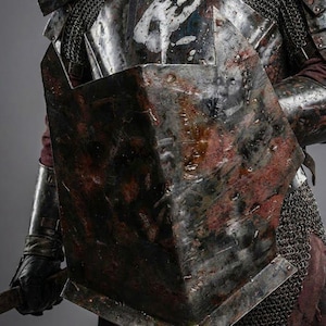 May include: Close-up of a medieval suit of armor, featuring a chest plate with a white handprint design and a shield. The armor has a weathered, battle-worn appearance with rust-colored accents. Chainmail and leather gloves are also visible.