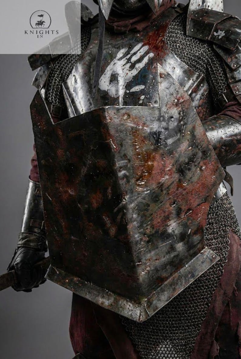 May include: Close-up of a medieval suit of armor, featuring a chest plate with a white handprint design and a shield. The armor has a weathered, battle-worn appearance with rust-colored accents. Chainmail and leather gloves are also visible.