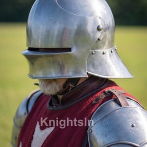 May include: A polished steel helmet with a curved top and visor, part of a medieval knight's armor. The helmet is riveted and worn over a red tunic with a white cross emblem. The image includes the text "Knights In".
