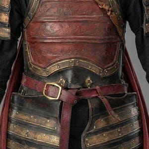 May include: Close-up of a medieval-style chest armor. The armor is primarily black with bronze-colored accents and a red leather belt. The chest plate has embossed detailing and is secured with a buckle. The Knights In logo is visible.