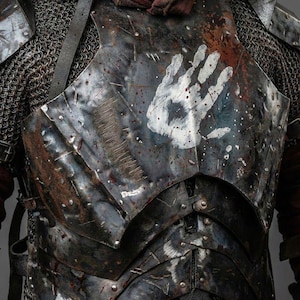 May include: Close-up of a weathered, dark metal breastplate with a white handprint. The armor shows signs of wear and tear, with rivets and layered plates. Chainmail is visible at the edges. The image includes the text "KNIGHTS IN".