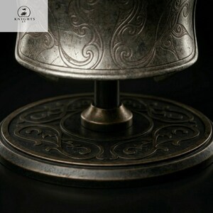 May include: Close-up of a metallic, antique-style object with intricate swirling patterns. The object appears to be a decorative piece, possibly a goblet or a stand, with a dark, aged finish. The base is circular with detailed engravings.