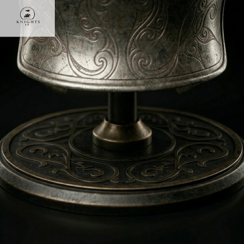 May include: Close-up of a metallic, antique-style object with intricate swirling patterns. The object appears to be a decorative piece, possibly a goblet or a stand, with a dark, aged finish. The base is circular with detailed engravings.