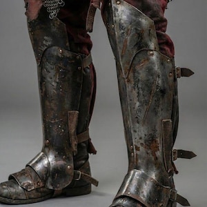 May include: Close-up of antique metal leg armor, featuring knee and shin protection. The armor is riveted and buckled, with a distressed, aged appearance. The image includes the text "KNIGHTS IN".
