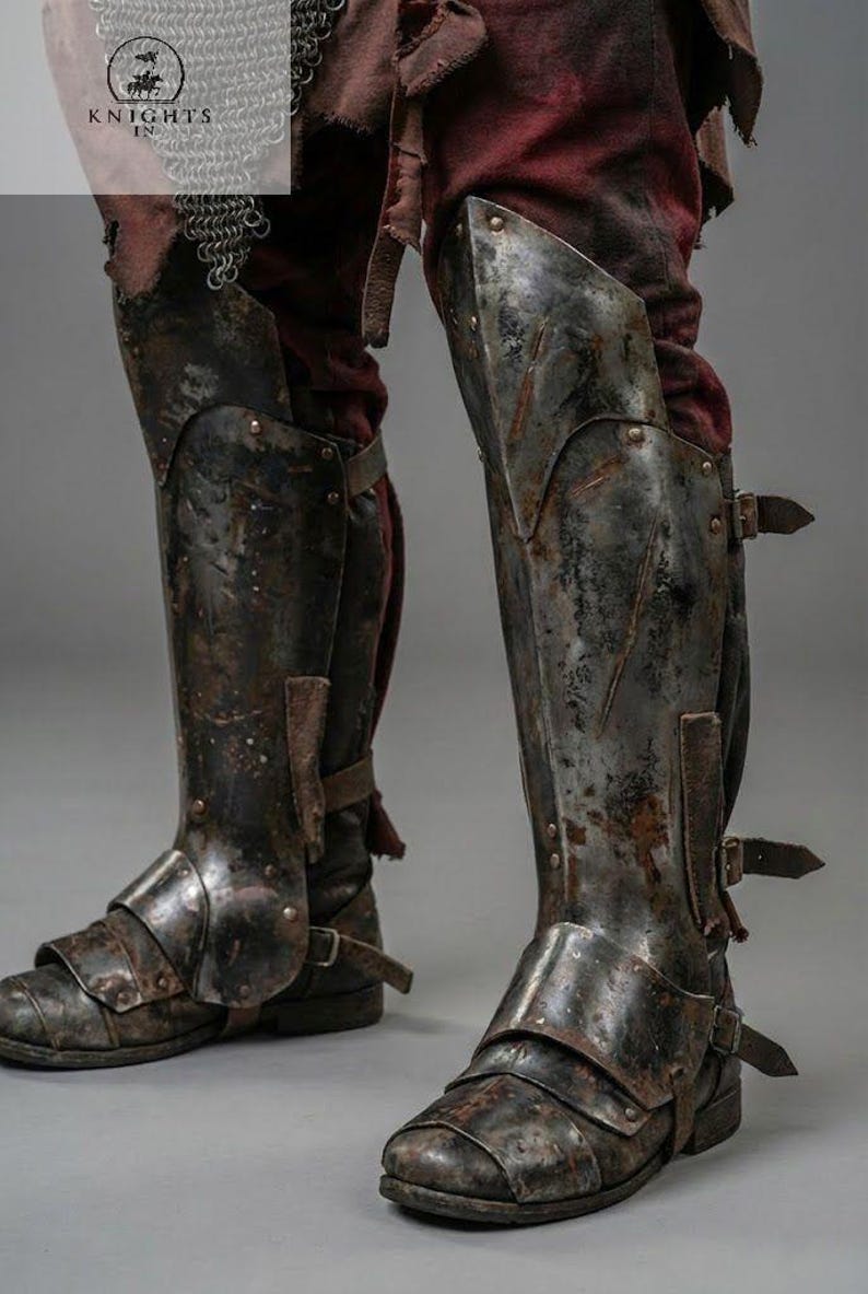 May include: Close-up of antique metal leg armor, featuring knee and shin protection. The armor is riveted and buckled, with a distressed, aged appearance. The image includes the text "KNIGHTS IN".