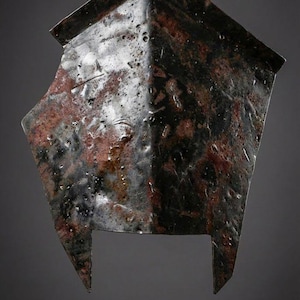 May include: A weathered, metal helmet with a dark, mottled finish. The helmet has a square top and two pointed extensions at the bottom. The surface shows signs of age and wear, with a mix of dark and reddish-brown tones. The logo "KNIGHTS IN" is in the upper left corner.