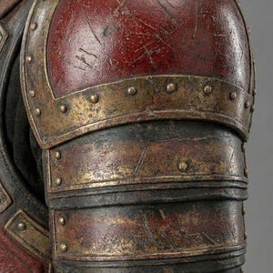 May include: Close-up of a weathered, antique-style shoulder armor piece. The armor features a deep red, slightly scratched surface with brass-colored accents and rivets. The image includes the text "KNIGHTS IN".