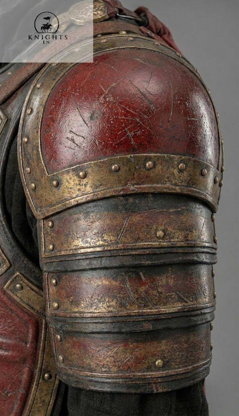 May include: Close-up of a weathered, antique-style shoulder armor piece. The armor features a deep red, slightly scratched surface with brass-colored accents and rivets. The image includes the text "KNIGHTS IN".