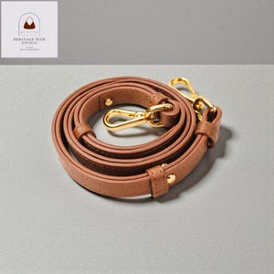May include: A brown leather bag strap with gold-colored metal clasps and hardware. The strap is coiled and ready for use. The Heritage Hide Studio logo is in the upper left corner.