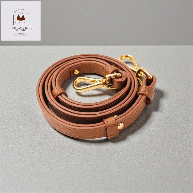 May include: A brown leather bag strap with gold-colored metal clasps and hardware. The strap is coiled and ready for use. The Heritage Hide Studio logo is in the upper left corner.