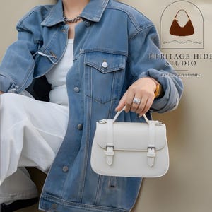 Vintage genuine leather crossbody handbag with retro Cambridge satchel design displayed in elegant neutral studio setting. Women’s minimalist shoulder purse available in black, burgundy red, ivory white, and blush pink colors with structured silhouette, buckle strap details, top handle, and adjustable crossbody strap. Japanese style leather handbag styled for modern fashion lovers, classic British satchel aesthetic, everyday women’s purse, elegant casual shoulder bag