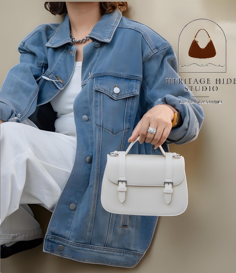 Vintage genuine leather crossbody handbag with retro Cambridge satchel design displayed in elegant neutral studio setting. Women’s minimalist shoulder purse available in black, burgundy red, ivory white, and blush pink colors with structured silhouette, buckle strap details, top handle, and adjustable crossbody strap. Japanese style leather handbag styled for modern fashion lovers, classic British satchel aesthetic, everyday women’s purse, elegant casual shoulder bag