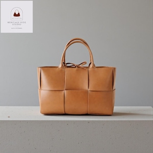 May include: A tan leather tote bag with a woven design on the front. The bag has two handles and a small bow detail at the top. The bag is sitting on a white surface. The logo "HERITAGE HIDE STUDIO" is in the top left corner.