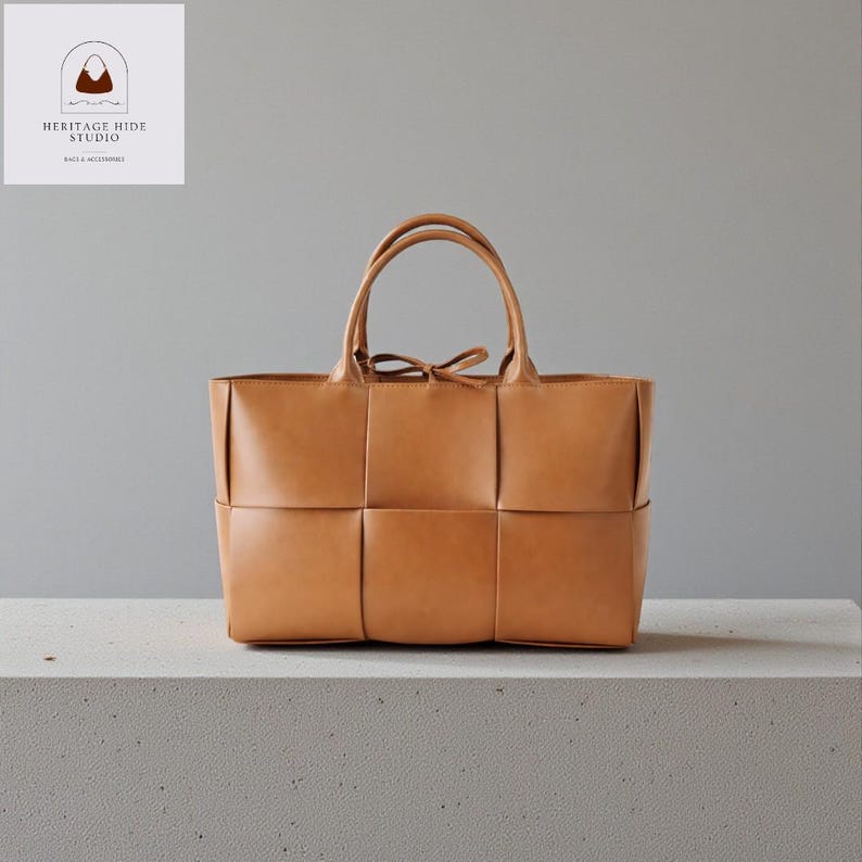 May include: A tan leather tote bag with a woven design on the front. The bag has two handles and a small bow detail at the top. The bag is sitting on a white surface. The logo "HERITAGE HIDE STUDIO" is in the top left corner.