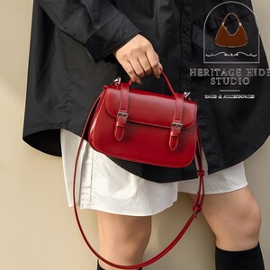 Vintage genuine leather crossbody handbag with retro Cambridge satchel design displayed in elegant neutral studio setting. Women’s minimalist shoulder purse available in black, burgundy red, ivory white, and blush pink colors with structured silhouette, buckle strap details, top handle, and adjustable crossbody strap. Japanese style leather handbag styled for modern fashion lovers, classic British satchel aesthetic, everyday women’s purse, elegant casual shoulder bag