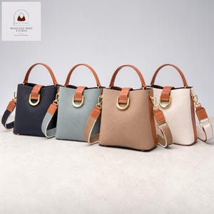 Puede incluir: Cuatro bolsos tipo cubo en azul marino, verde salvia, beige y blanco roto. Cada bolso presenta un asa y detalles de correa marrones, una anilla en D dorada y una correa de hombro a rayas desmontable. Los bolsos se exhiben sobre un fondo neutro.