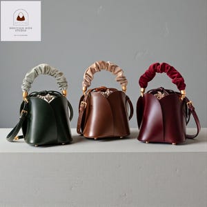 May include: Three leather bucket bags in dark green, brown, and burgundy. Each bag has a gathered velvet handle in coordinating colours, gold hardware, and a drawstring closure. The bags are displayed on a neutral surface.
