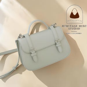 Vintage genuine leather crossbody handbag with retro Cambridge satchel design displayed in elegant neutral studio setting. Women’s minimalist shoulder purse available in black, burgundy red, ivory white, and blush pink colors with structured silhouette, buckle strap details, top handle, and adjustable crossbody strap. Japanese style leather handbag styled for modern fashion lovers, classic British satchel aesthetic, everyday women’s purse, elegant casual shoulder bag