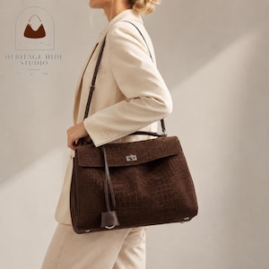 May include: A dark brown leather handbag with a textured crocodile-embossed pattern, featuring a rectangular flap, a silver-toned metal turn-lock closure, a long shoulder strap, and a dangling leather charm, worn with a cream-colored blazer.