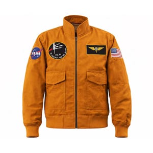 Könnte beinhalten: Senffarbene Bomberjacke mit Stehkragen, zwei Vordertaschen und Reißverschluss. Mit NASA- und US Air Force-Patches sowie einem amerikanischen Flaggen-Patch am Ärmel. Die Jacke hat gerippte Bündchen und einen gerippten Saum.