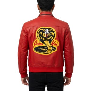 May include: A red leather jacket featuring a large embroidered Cobra Kai logo on the back. The logo depicts a black and yellow cobra snake with the text Cobra Kai in yellow script. The jacket has a stand-up collar, ribbed cuffs, and a ribbed hem.
