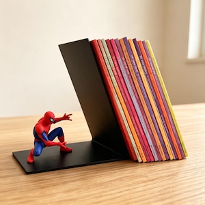 以下が含まれることがあります： 黒い金属製のブックエンドで、赤と青のスパイダーマンのフィギュアがしゃがんだポーズをとっています。ブックエンドは、背表紙に様々なタイトルが印刷された12冊の薄くてカラフルな本を支えており、斜めに並べられています。