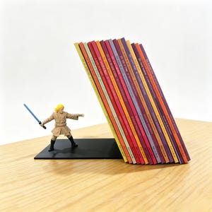Peut inclure: Un serre-livres en métal noir avec une figurine miniature d'un personnage blond en robe beige tenant un sabre laser bleu. Le serre-livres soutient une rangée de quinze livres fins et colorés aux dos marqués Arcturus, inclinés en pile.