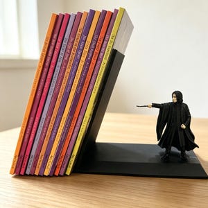 Può includere: Un fermalibri nero a forma di L con una miniatura di un personaggio in una lunga veste nera che tiene una bacchetta. Il fermalibri sostiene una fila di dodici libri tascabili colorati di Arthur Conan Doyle, ognuno con un colore del dorso distinto.