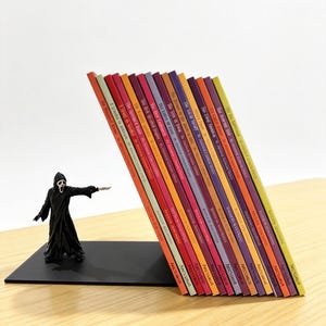 Peut inclure: Un serre-livres en métal noir représentant une figurine Ghostface tenant un couteau, soutenant une rangée de quinze livres colorés avec des titres en blanc sur les tranches. Les livres sont inclinés contre la figurine sur une base noire.