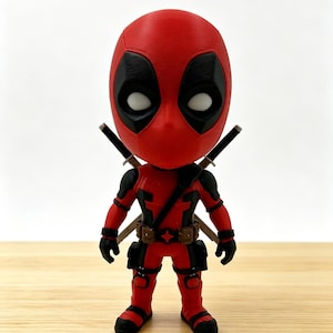 May include: A stylised figurine of a character with a large head, wearing a red and black suit with a utility belt featuring a star-shaped buckle. The figure has white eye patches, black accents on the limbs, and two swords crossed behind its back.