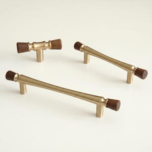 May include: Four cabinet pulls with a mid-century modern design. The pulls have a brass finish with dark brown wooden ends. Two are long, curved pulls, one is a shorter, straight pull, and the last is a T-shaped pull.