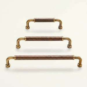 May include: Three cabinet pulls with a brass finish and dark wood center sections. The pulls are arranged vertically, showcasing variations in size. The design features a curved shape with a classic, elegant aesthetic.
