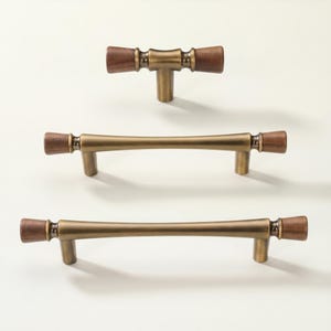 Könnte beinhalten: Drei Schrankgriffe im Mid-Century-Modern-Design. Die Griffe haben eine Messingoberfläche mit Holzakzenten an den Enden. Der obere Griff ist T-förmig, die anderen beiden sind stangenförmig.
