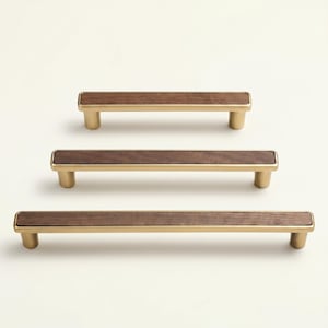 May include: Three cabinet pulls with a rectangular walnut wood center and brass-colored metal handles. The pulls are arranged vertically, showcasing variations in size. The wood grain is visible, adding a natural element to the design.