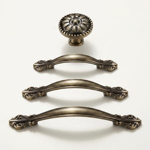 May include: An assortment of antique bronze cabinet hardware, including a round knob with a floral design and three matching pull handles. The hardware is displayed on a white surface.