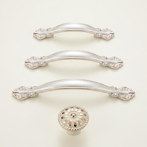 May include: A set of silver-toned cabinet hardware, including three arched handles and a round knob. The handles have decorative ends, and the knob features a floral design with gold accents. The hardware is designed for furniture.