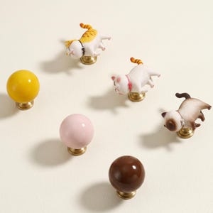 May include: A collection of decorative cabinet knobs. The knobs include yellow, pink, and brown spherical designs, along with cat-shaped knobs in various colors. Each knob has a brass base, creating a charming and whimsical aesthetic.
