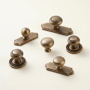 May include: An assortment of antique brass cabinet knobs and pulls. The knobs have various designs, including a round ball, a rope-like texture, and a stepped design. The pulls have a rectangular backplate with a decorative edge.