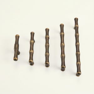 May include: A collection of five bronze-colored bamboo-style cabinet pulls of varying lengths. Each pull features a cylindrical design with segmented detailing, mimicking bamboo stalks. The hardware is set against a plain white background.