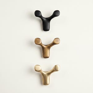Puede incluir: Tres perchas de pared en fila. La percha superior es negra, la del medio es bronce y la inferior es dorada. Cada percha tiene un diseño en forma de Y con extremos redondeados, adecuado para colgar objetos.