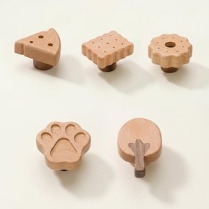 May include: A collection of five wooden cabinet knobs in various shapes: a cheese wedge, a cracker, a biscuit, a paw print, and a tree. Each knob has a light brown finish with a dark brown base or accent.