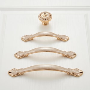 May include: A set of decorative cabinet hardware featuring a gold finish. The set includes a round knob and three matching pull handles. The handles have a cream-colored center and ornate detailing on the ends, suitable for drawers or cabinets.