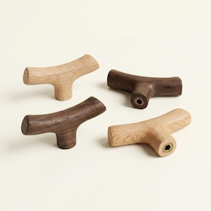 Puede incluir: Cuatro tiradores de madera para armarios en tonos marrones y beige. Los tiradores tienen un diseño en forma de T, con un mango redondeado y una base plana. Dos son de color claro y dos de color oscuro, mostrando la veta natural de la madera.
