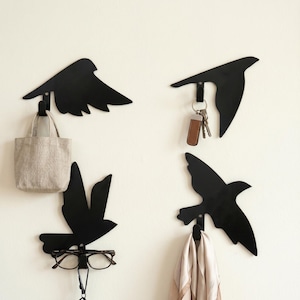 Può includere: Quattro ganci da parete neri a forma di uccello. Ogni gancio è una diversa silhouette di uccello, con un gancio sotto. Un gancio tiene una piccola borsa tote beige, un altro chiavi, un terzo occhiali e l'ultimo una sciarpa.