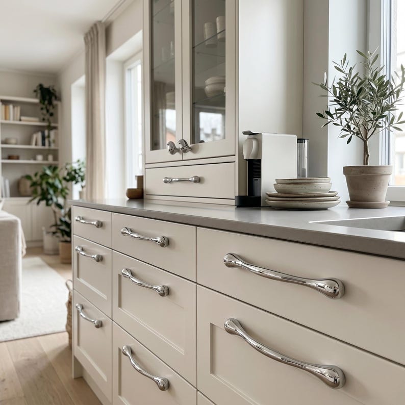 Puede incluir: Una cocina moderna con gabinetes de color beige claro y tiradores de cajones cromados pulidos. Un peque&ntilde;o olivo en una maceta de terracota se encuentra en la encimera cerca de una ventana. Tambi&eacute;n se ve una cafetera y platos apilados.